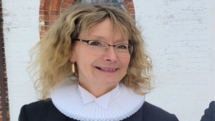 En person iført briller og formelt tøj, herunder en hvid flip, der står nær et buet murstensvindue.