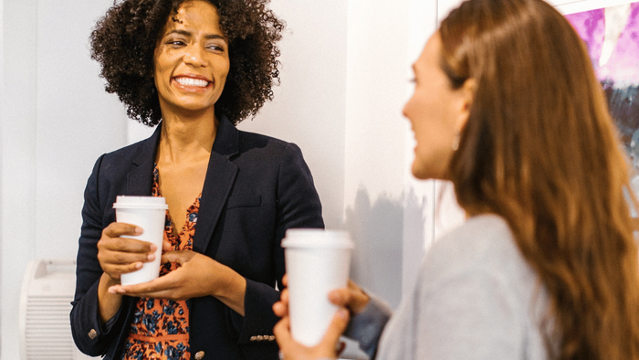 Zwei Frauen in professioneller Kleidung, die in einem sozialen oder geschäftlichen Umfeld lächelnd Kaffeetassen halten.