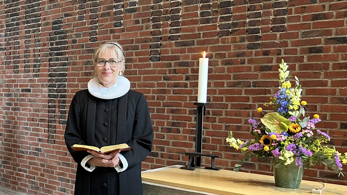 En kvinde i svart kapittel holder en bog og står foran en mur af røde mursten.