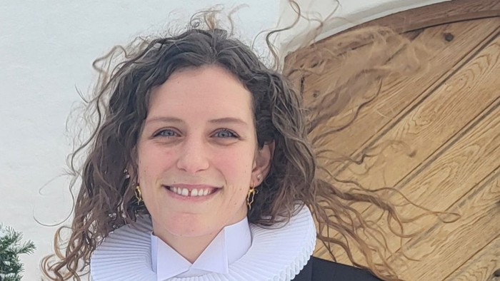 Person med et smil iført akademisk kappe med hvid krave og guldøreringe udendørs.