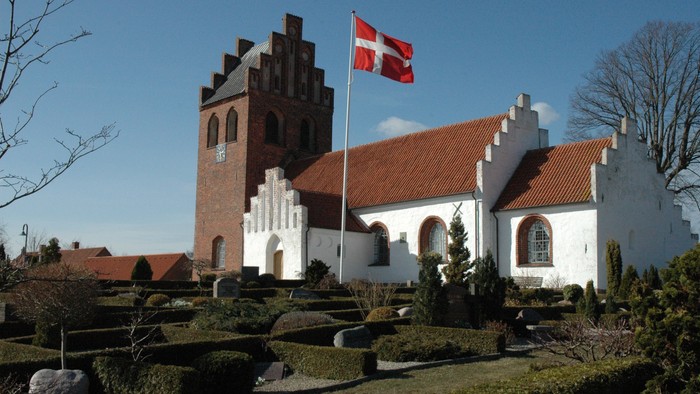 En malerisk kirke med et dansk flag vajende på toppen, omgivet af velplejede haver og træer.