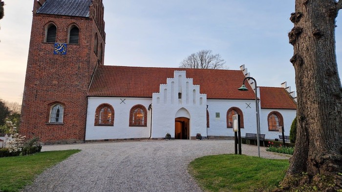 Historisk kirke med murstens tårn og hvid facade, placeret på en grussti omgivet af træer.