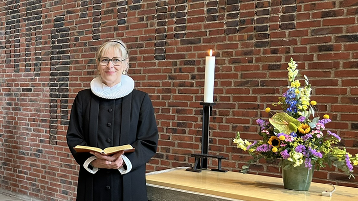 En kvinde i svart kapittel holder en bog og står foran en mur af røde mursten.