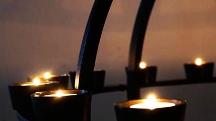 Flere tændte stearinlys i mørke lysestager mod en lys væg.