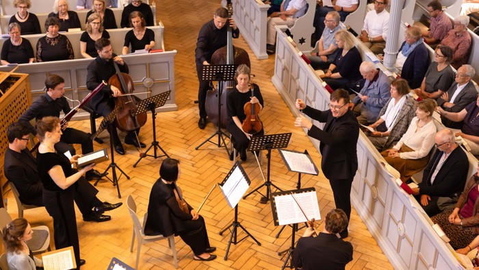 Orchester, das in einer Kirche mit Publikum in den Kirchenbänken auftritt, dirigiert von einem Dirigenten.