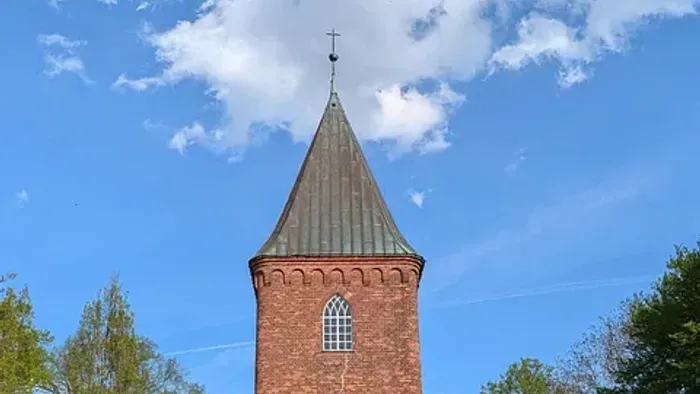 Kirchturm aus rotem Backstein vor blauem Himmel und Bäumen