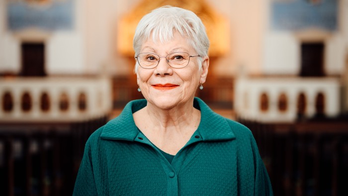 En ældre dame med kort, hvidt hår og briller, iført en grøn skjorte, smiler varmt.