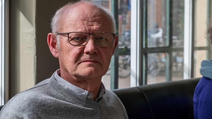 En skaldet mand med briller iført en grå sweater kigger tankefuldt ud ad et vindue.