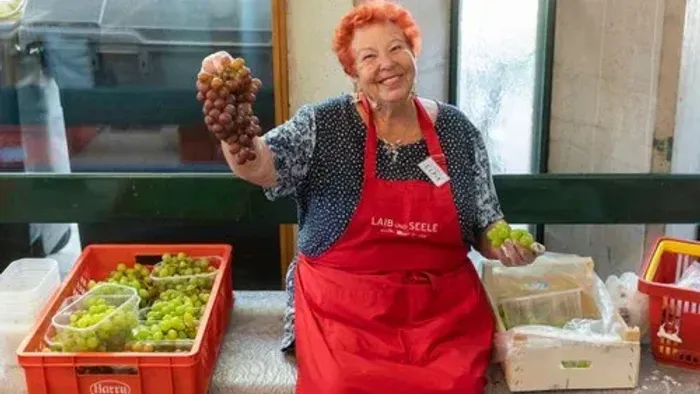 Eine Frau in einer roten Schürze hält eine Traube hoch und steht vor Kisten mit Obst.