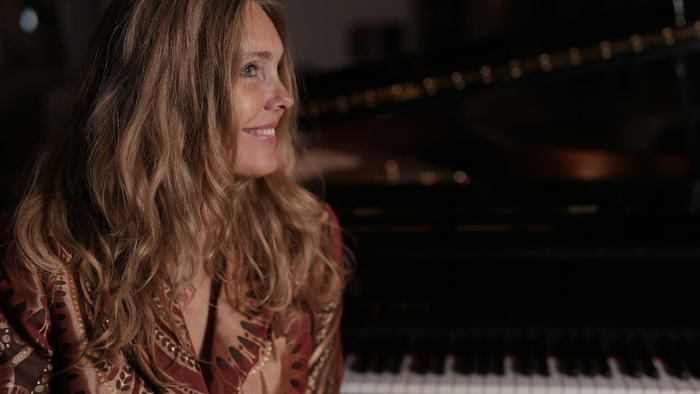 En kvinde med langt, blond hår sitter ved et piano og smiler.