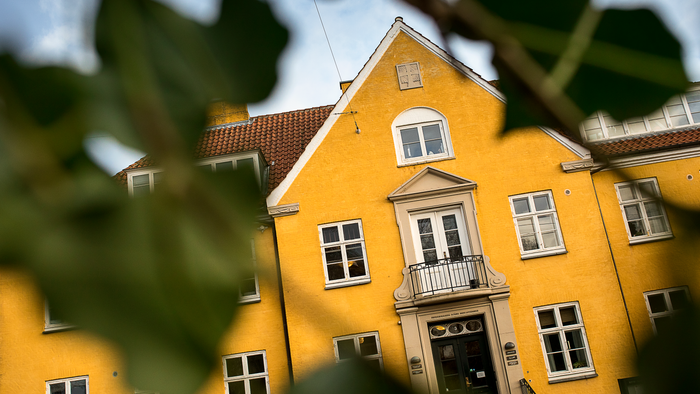 Lysende gul bygning med hvidt udsmykning, delvist skjult af grønt løv, med klassiske arkitektoniske detaljer.