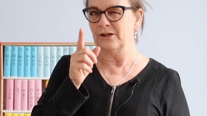 En kvinde med briller gestikulerer, mens hun står foran en farverig bogreol.