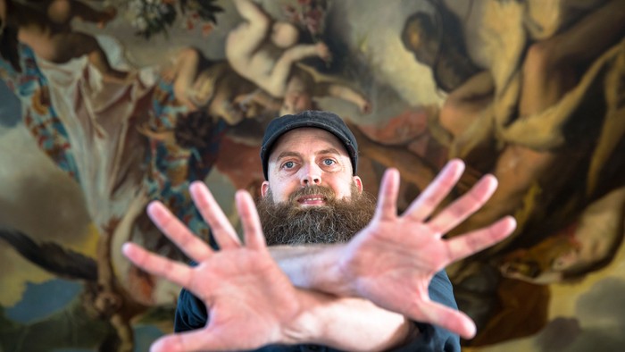 Mand med skæg og mørk behøjning har hænderne op foran maleri