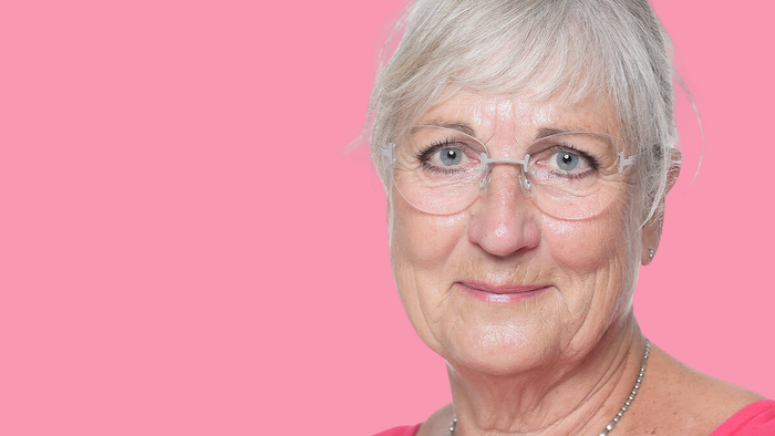 En smilende ældre kvinde med kort gråt hår, briller og et perlekæde på en lyserød baggrund.
