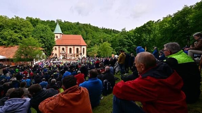 Eine große Menschenmenge versammelt sich im Freien nahe einer Kirche in einer grünen, hügeligen Gegend für eine Veranstaltung.