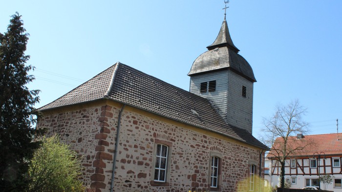 Steinkirche mit Glockenturm und Kreuz, umgeben von Grünflächen.
