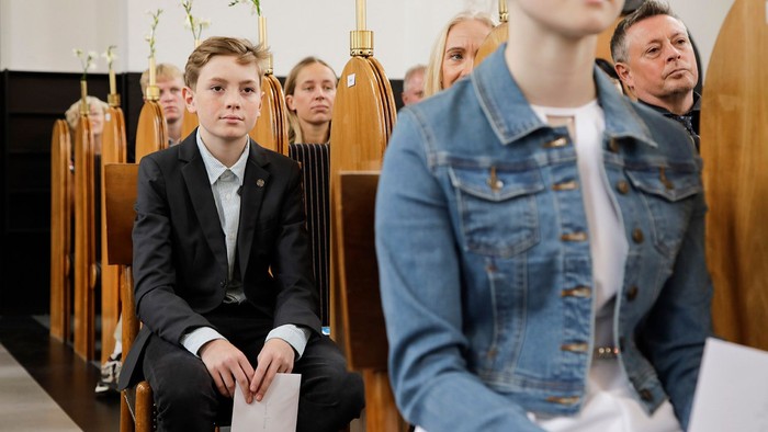 En højtidelig forsamling i en kirke med opmærksomme deltagere, herunder en ung dreng i pæn påklædning.