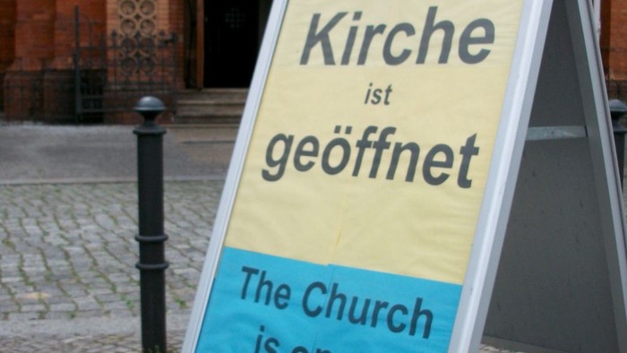 Vor einer Kirche steht ein zweisprachiges Schild mit der Aufschrift *„The Church is open“* auf Englisch und *„Die Kirche ist geöffnet“* auf Deutsch.