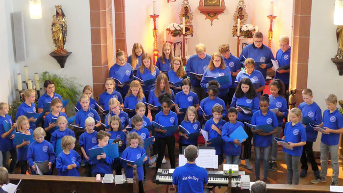 Ein Kinderchor tritt in einer Kirche auf, trägt blaue Hemden und hält Gesangbücher in den Händen.