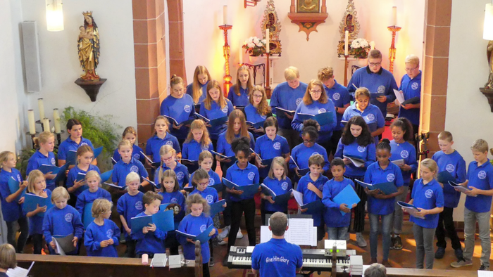 Ein Kinderchor tritt in einer Kirche auf, trägt blaue Hemden und hält Gesangbücher in den Händen.