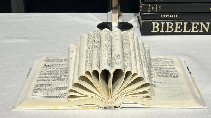 Bibel ligger åbnet på et alterbord - en del af siderne er foldet indad og danner en vifte. En anden bibel, en ritualbog og andre bøger i baggrunden