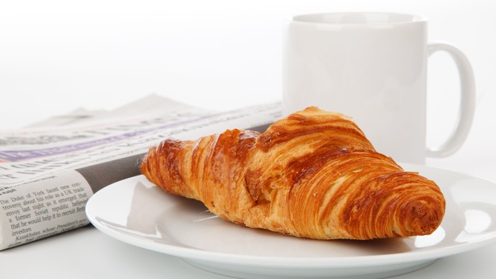 Ein Croissant auf einem Teller neben einer Tasse und einer Zeitung