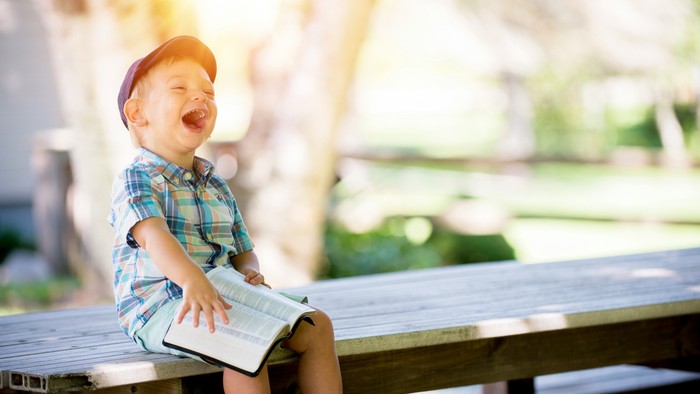 A young boy in a plaid shirt and baseball cap sits on a park bench, laughing while holding a book.