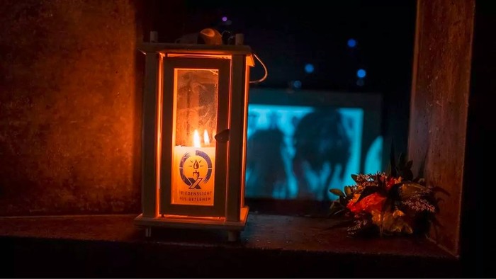 Eine erleuchtete Laterne auf einer Fensterbank mit einem dekorativen Kranz daneben, die auf eine dunkle Nacht hinausblickt.