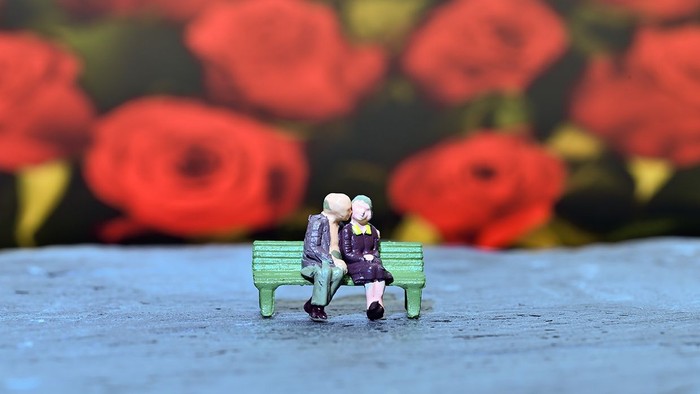 Winzige Figuren sitzen liebevoll auf einer Parkbank, umgeben von leuchtend roten Blumen.