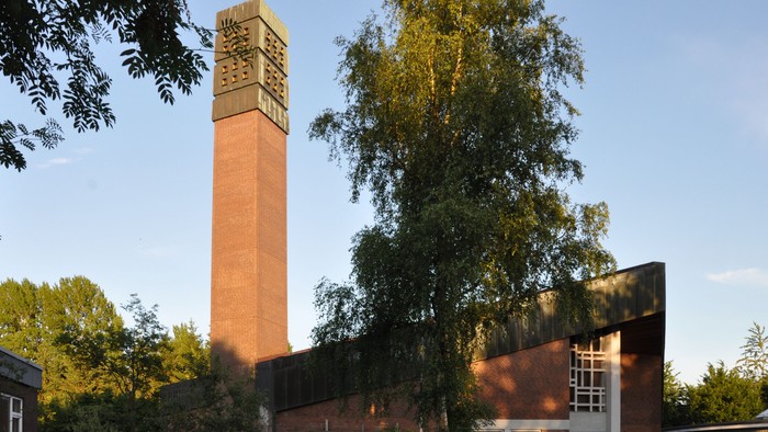 Ein roter Ziegelbau mit hohem Turm und Uhr, umgeben von Bäumen und Gras.