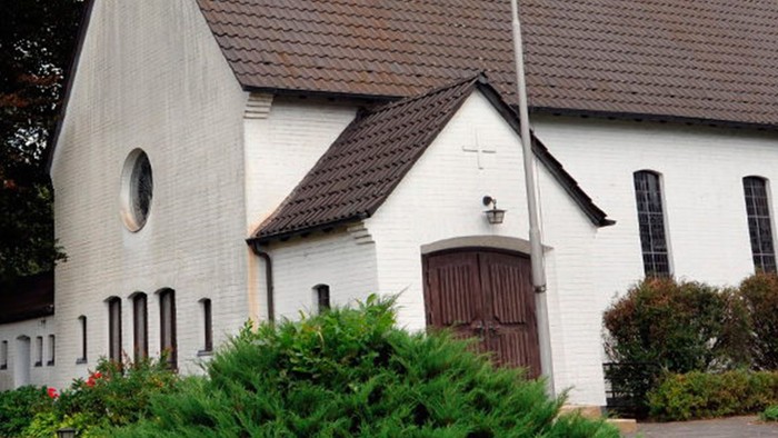 Weiße Kirche mit hohem Turm und braunem Dach, umgeben von Bäumen und Büschen