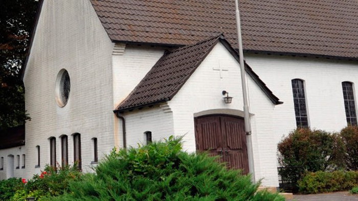 Weiße Kirche mit hohem Turm und braunem Dach, umgeben von Bäumen und Büschen