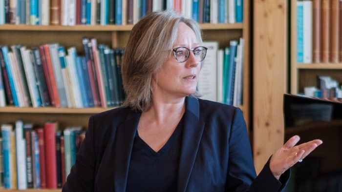 Kvinde i blå jakke og briller sidder i bibliotek og holder pen i hånden