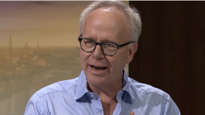 En mand med briller og en lyseblå skjorte gestikulerer, mens han taler under en samtale.