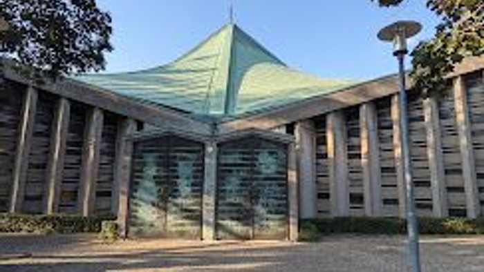 Modernes Gebäude mit grünem Dach und großen Glasfenstern, umgeben von Bäumen und einem Kopfsteinpflasterbereich.