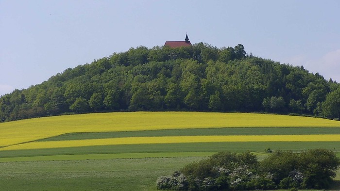 Üppiger grüner Hügel mit einer Kapelle, gekrönt von leuchtend gelben Feldern und rotgedeckten Häusern darunter.