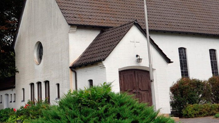 Weiße Kirche mit hohem Turm und braunem Dach, umgeben von Bäumen und Büschen