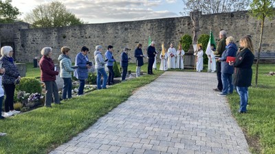Die Gruppe versammelt sich bei einer Grabzeremonie, während Geistliche Fahnen und Blumen in einer feierlichen Atmosphäre im Freien halten.