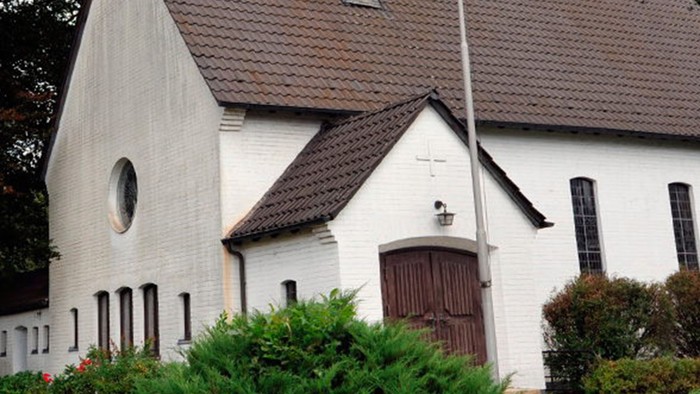 Weiße Kirche mit hohem Turm und braunem Dach, umgeben von Bäumen und Büschen