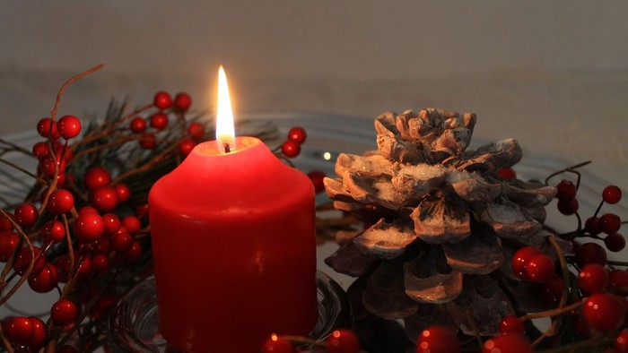 Eine rote Kerze brennt neben einem Tannenzapfen und roten Beeren