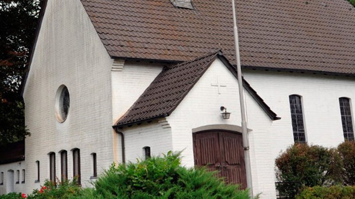 Weiße Kirche mit hohem Turm und braunem Dach, umgeben von Bäumen und Büschen