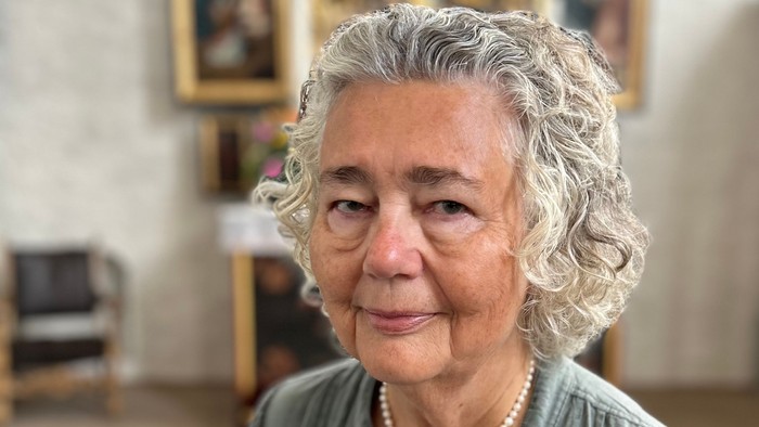 Ældre dame med krøllet gråt hår, der bærer en perlehalskæde, og står i en kirke.