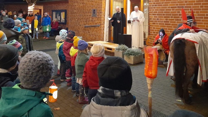 Viele Kinder und Erwachsene stehen mit Laternen vor der Kirche, während Pastor Fitschen und Gemeindereferent Becker die Martinsandacht halten. An der rechten Bildseite sind der Bettler und St. Martin neben seinem Pferd zu sehen.