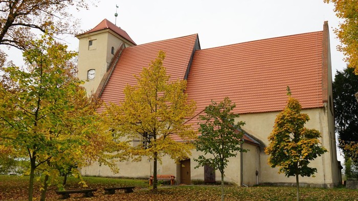 Kirche mit rotem Satteldach und Turm, umgeben von herbstlich gefärbten Bäumen auf einer Wiese.