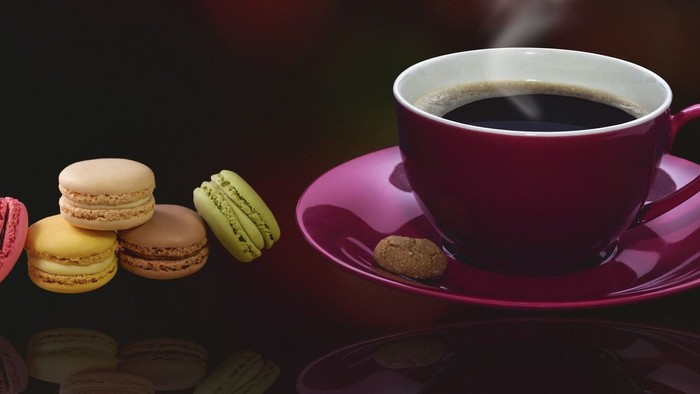 Ein pinker Tassen mit dampfendem Kaffee und bunte Macarons auf einem schwarzen Untersetzer