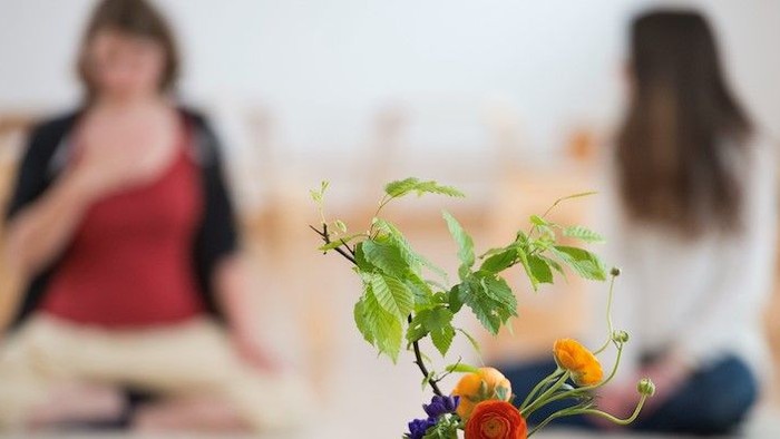Menschen meditieren in einer ruhigen Umgebung mit einer Vase voller bunter Blumen im Vordergrund.