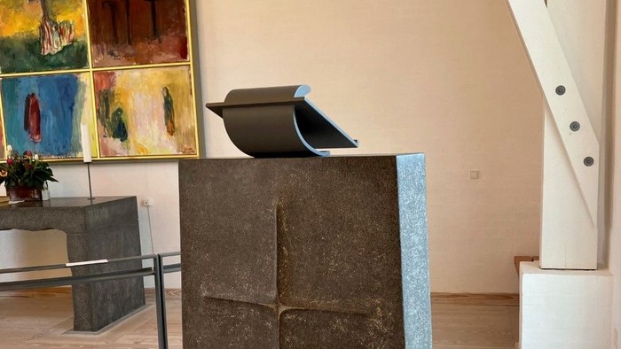 En stor sten med et udhugget kors står i et rum med billeder og trappe.