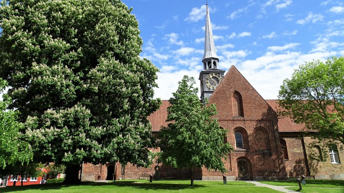 En rød murstenskirke med tårn og klokke, omgivet af grønne træer og en græsplæne