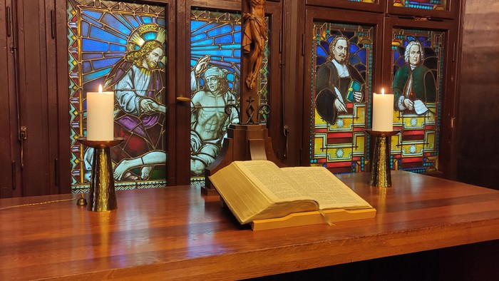 Der Altar im Gemeindehaus Kreuz vor bunten Glasscheiben. Darauf zwei leuchtende Kerzen und eine aufgeschlagene Bibel.
