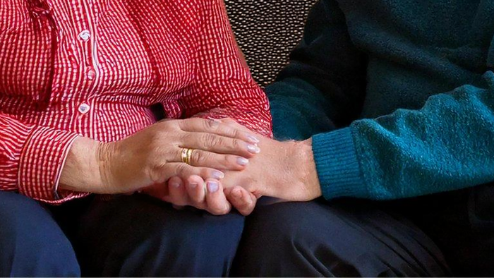 Zwei Personen, die sich an den Händen halten: eine trägt ein rotes kariertes Hemd, die andere einen blauen Pullover.